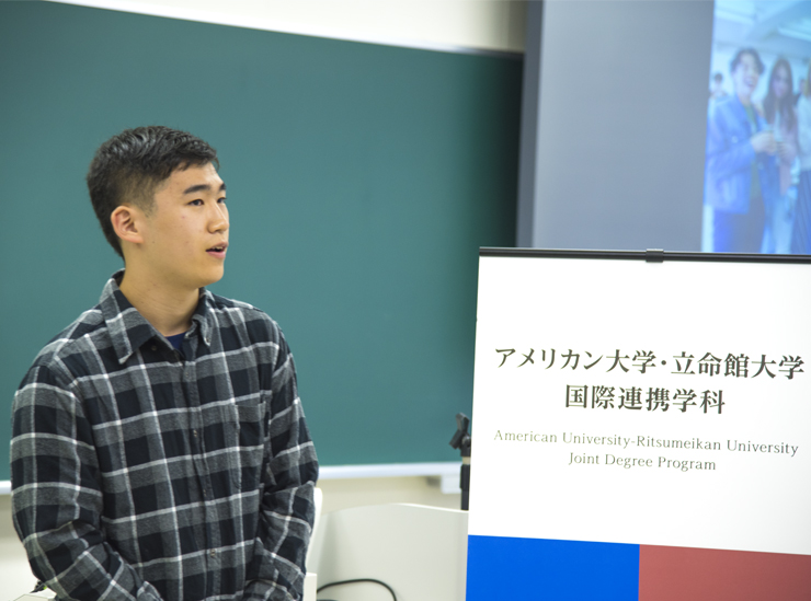 A Joint Degree Program student stands in a classroom giving his informal speech wearing a check black white and grey shirt.