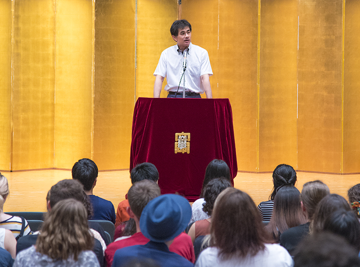 Associate Professor Takamichi Isoda, Director of the International Center, stands on stage at a lectern draped in a thick burgundy red cloth covering emblazoned with the traditional university icon
