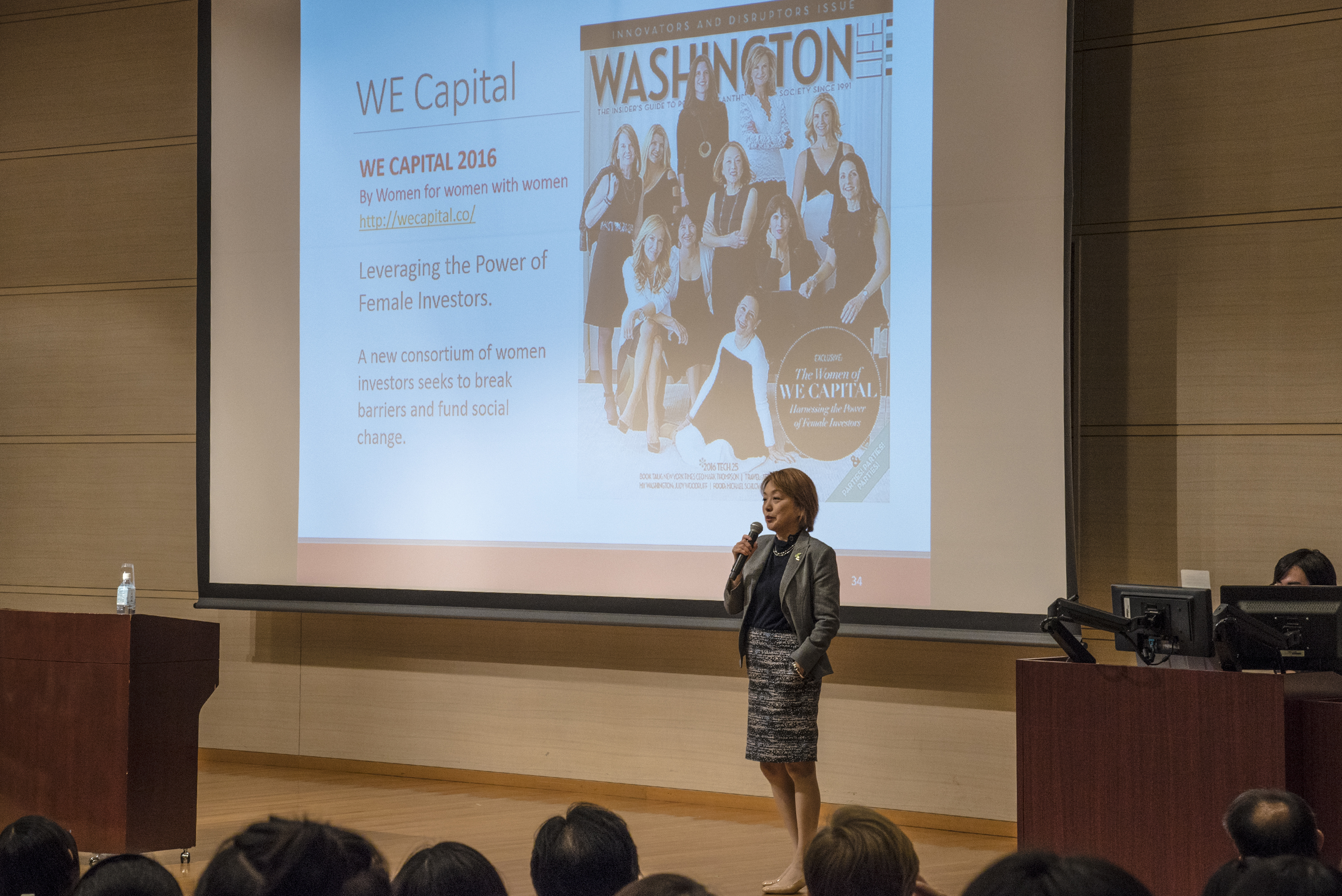 Dr. Kuno stands on stage. In the background is a slide of a front page of Washington magazine featuring Dr. Kuno