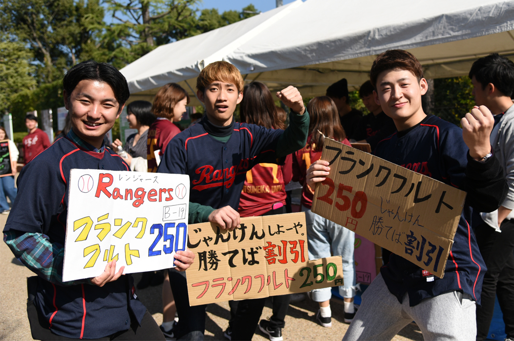 Members of the ‘Rangers’ rubber-ball baseball team with handwritten signs advertise Frankfurt sausages for 250 yen and a discount for customers who beat them at rock, paper scissors.