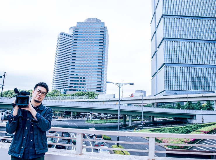 Director Takao Gotsu stands on a bridge over a busy multiple lane road, camera perched on shoulder 