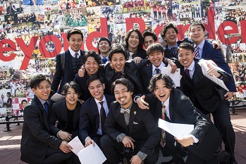 A group of students pose for a commemorative photo in front of the photo board outside on campus