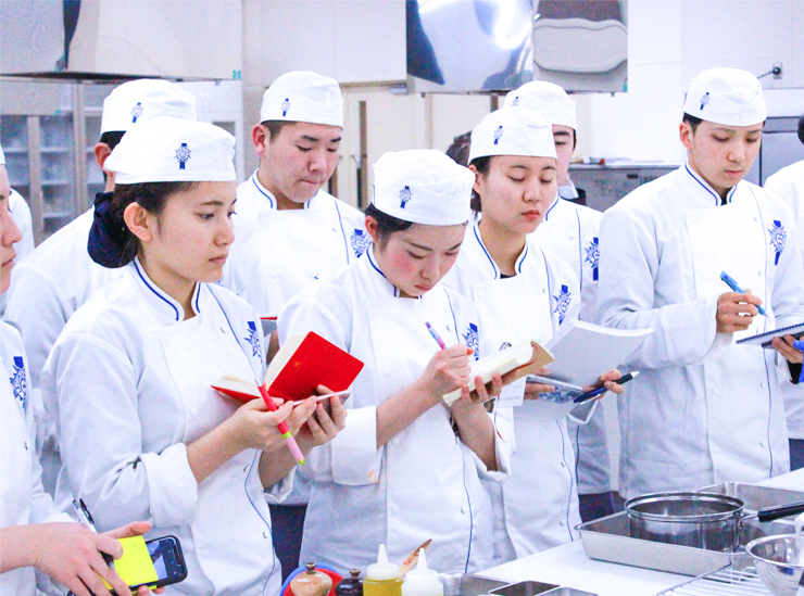 Ritsumeikan University students on the Global Culinary Arts and Management Programme take notes standing up wearing their chef's uniform
