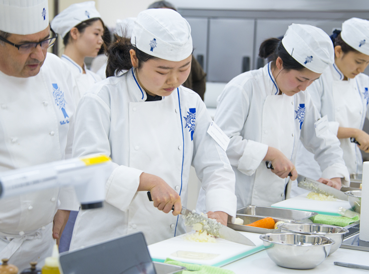 Ritsumeikan University College of Gastronomy Management students in chef whites concentrate intensely on chopping carrots and other vegetables to size under the watchful eye of Le Cordon Bleu Chef Gilles Company