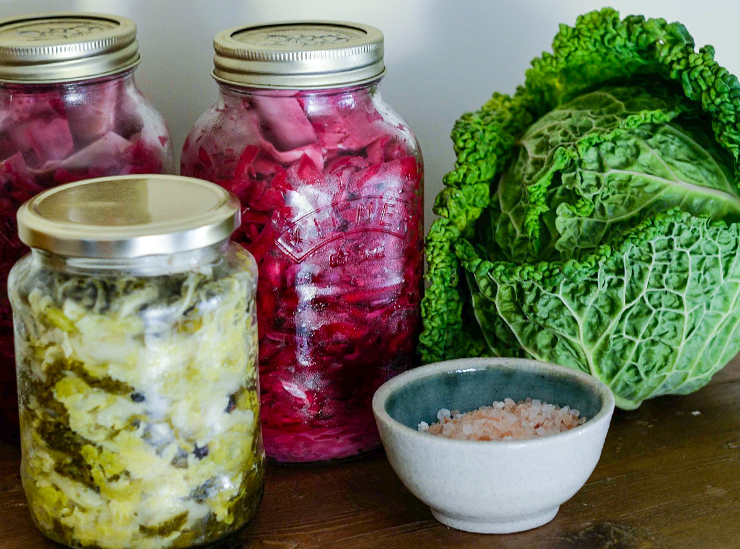 Three jars of homemade pickled cabbage, each a different color, sit on a table together with a small bowl of salt and a healthy-looking ripe green cabbage