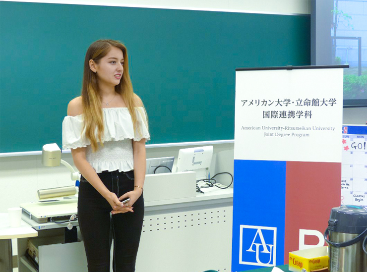A Joint Degree Program student stands in a classroom giving her informal speech wearing a white top and black trousers, phone in hand, by a sign with the logos of RU and AU