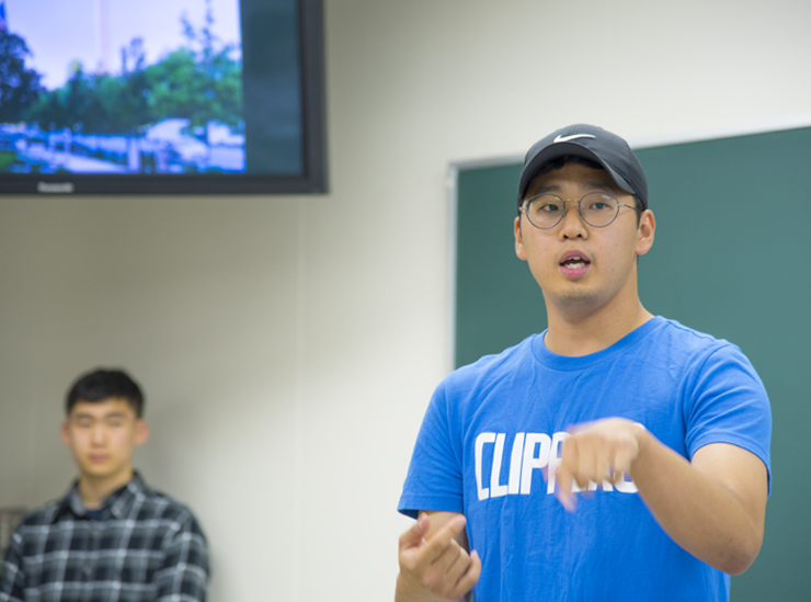 A Joint Degree Program student stands in a classroom giving his informal speech wearing a blue t-shirt, wearing a baseball cap, with a student looking on in the background