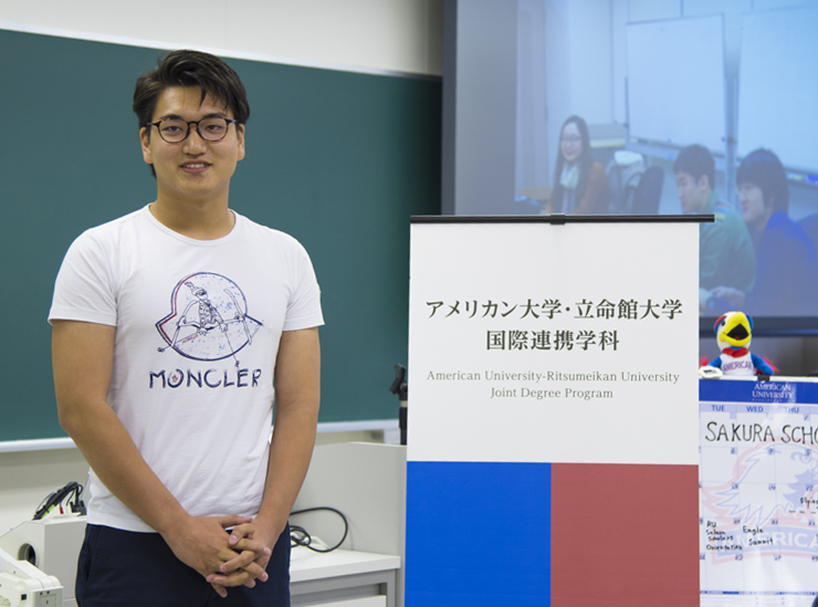 A Joint Degree Program student stands in a classroom giving his informal speech wearing a white t-shirt and black trousers, by a sign with the logos of RU and AU