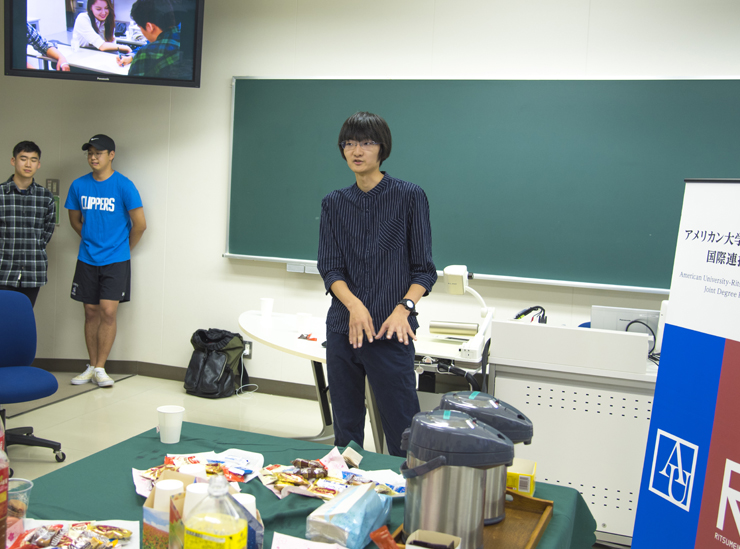 A Joint Degree Program student stands in a classroom giving his informal speech. The shot is zoomed out and so the table with refreshments on can be seen in the foreground. Two students listen in the background. The student himself is wearing a dark shirt with the sleeves rolled up and black trousers