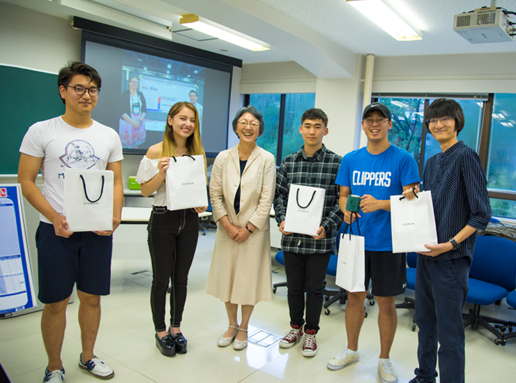 The five students stand with dean Kawamura. All are smiling and the students are holding their white send-off gift bags with black string handles and Ritsumeikan written across the front in small lettering