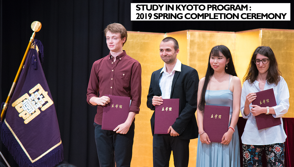 Four students of Ritsumeikan University's Study in Kyoto Program stand on stage, burgundy completion certificate binders under arm. To their left is the university flag. In the background is a Japanese gold-leaf gilded screen.
