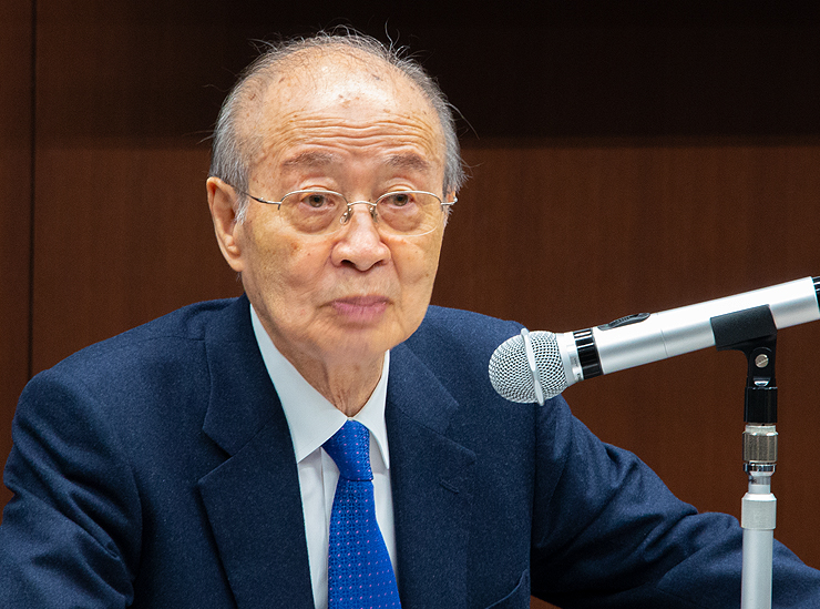Mr. Akashi at 89 years of age sits at a desk with a microphone wearing a smart blue suit replete with an immaculate square-dotted geometrically patterned slightly darker blue tie 